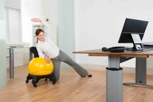 Young Happy Businesswoman Doing Fitness Exercise On Yellow Pilates Ball In Office